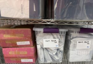 Shelf in the Valley Health Logistics storage unit holding pink Warm Hearth cartons and two clear bins hand-labeled Warm Hearth Tea 30, with bagged Yo Yi Patch inventory on the shelf above.